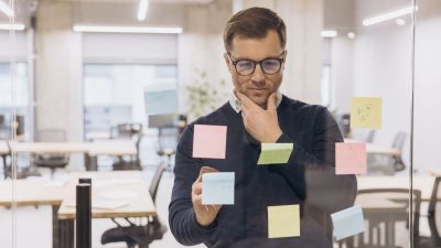 Projektleiter an Whiteboard schaut auf Notizen und ist in Gedanken. 