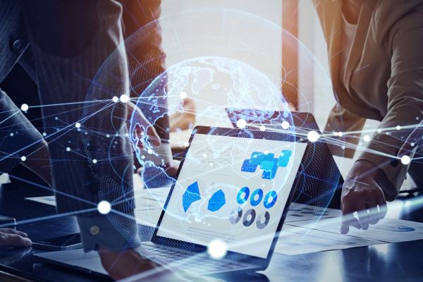 Laptop mit Daten-Dashboard auf einem Konferenztisch, umgeben von Geschäftsleuten, die Dokumente prüfen. Im Vordergrund ist eine digitale Weltkugel mit vernetzten Linien eingeblendet, Symbol für IT-Governance, globale Vernetzung und Datenanalyse. 
