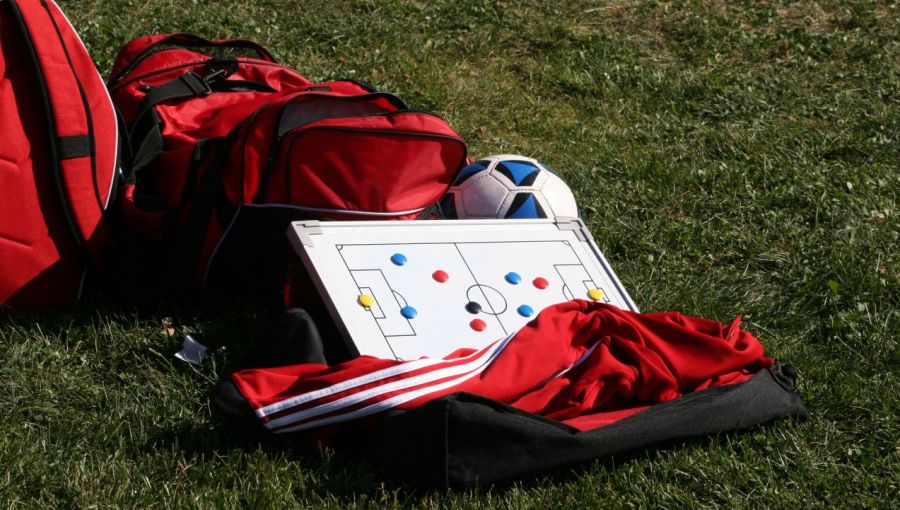 Rote Sporttaschen, Fußball und Taktiktafel mit Spielfeldmarkierung auf grünem Rasen: Symbol für Strategie, Planung und Teamarbeit im Projektmanagement.