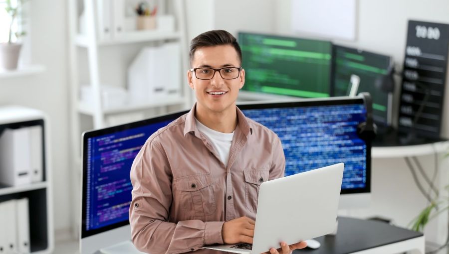 IT-Spezialist mit Laptop im modernen Büro vor Monitoren mit Programmiercode: Symbol für Personaldienstleistung und IT-Fachkräfte. 